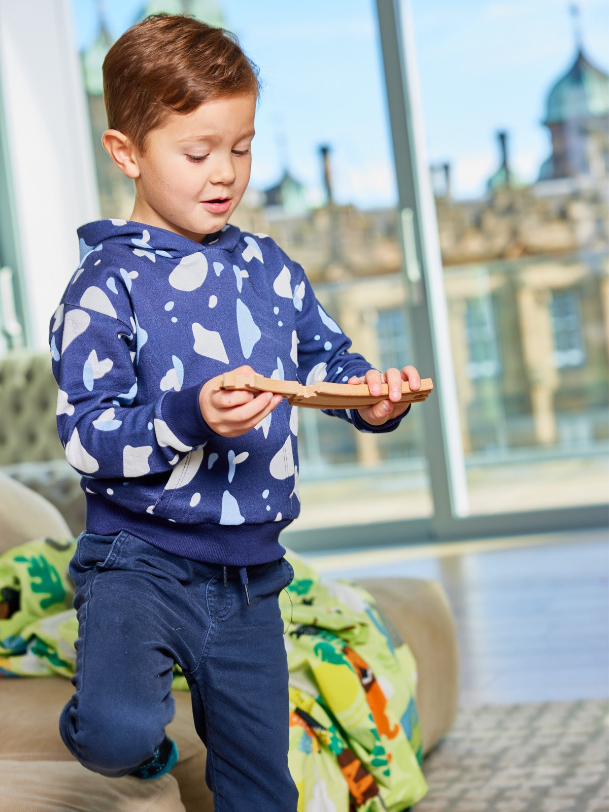 A child pictured playing with a wooden toy piece, wearing the Seb sustainable kids hoodie in navy with a blue and white terrazzo print.