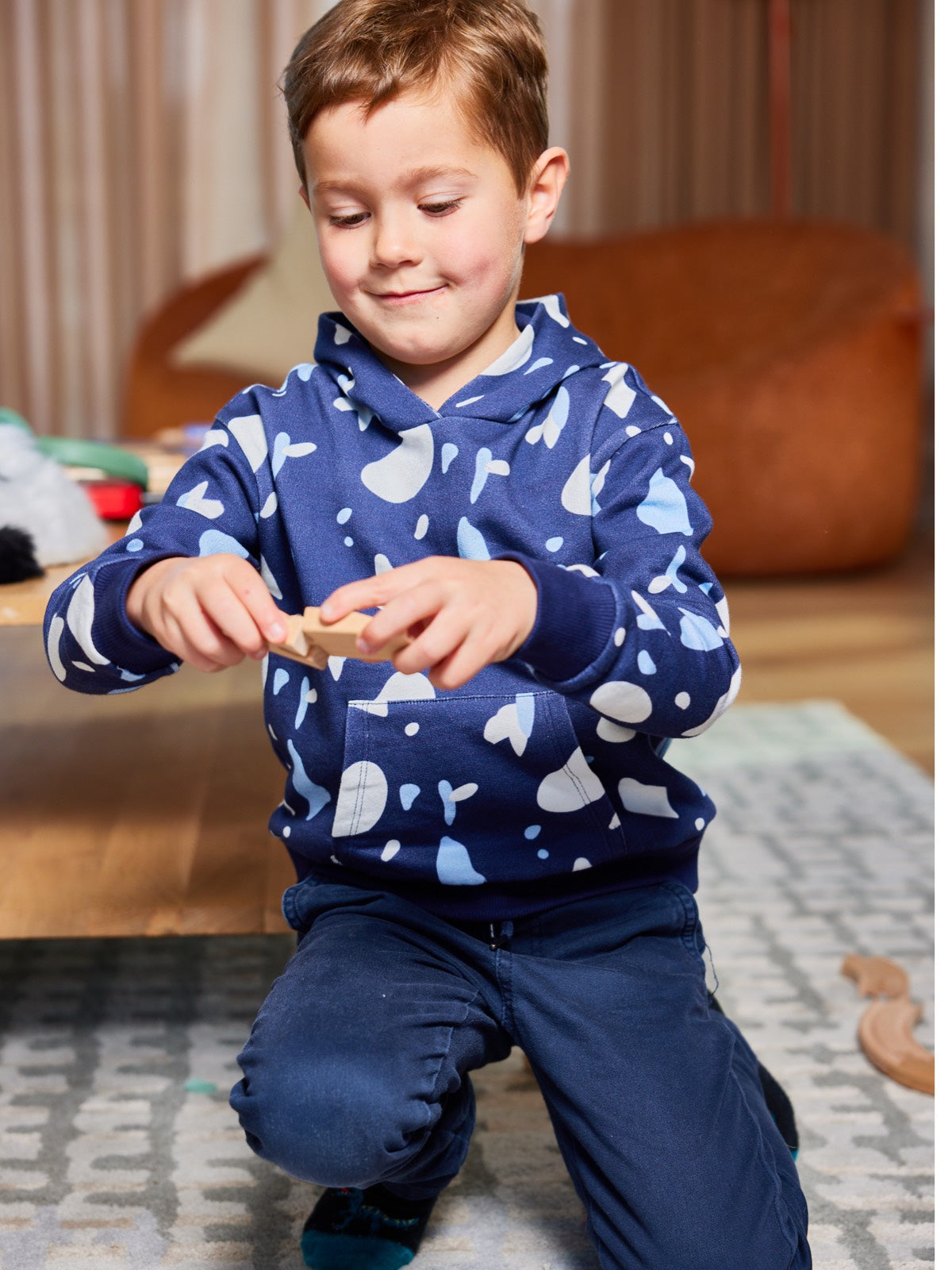 A child wearing the Seb sustainable kids hoodie in navy with a blue and white terrazzo print, pictured playing with a wooden toy piece and grinning. 