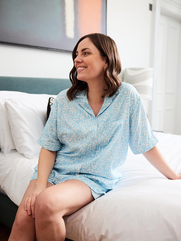 A model is pictured wearing the Sian Pyjamas in blue print whilst sat in a bedroom on the bed. There is artwork on the wall and the room has lots of natural light. 