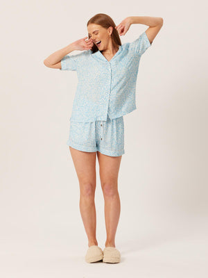 A model wears the Sian pyjama set in blue abstract leaf print. They are pictured wearing slippers and yawning and stretching.  