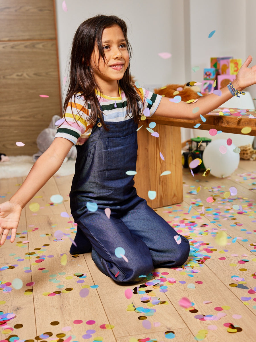 A child wearing the Sofia sustainable kids dungarees, pictured on the floor with falling confetti. 