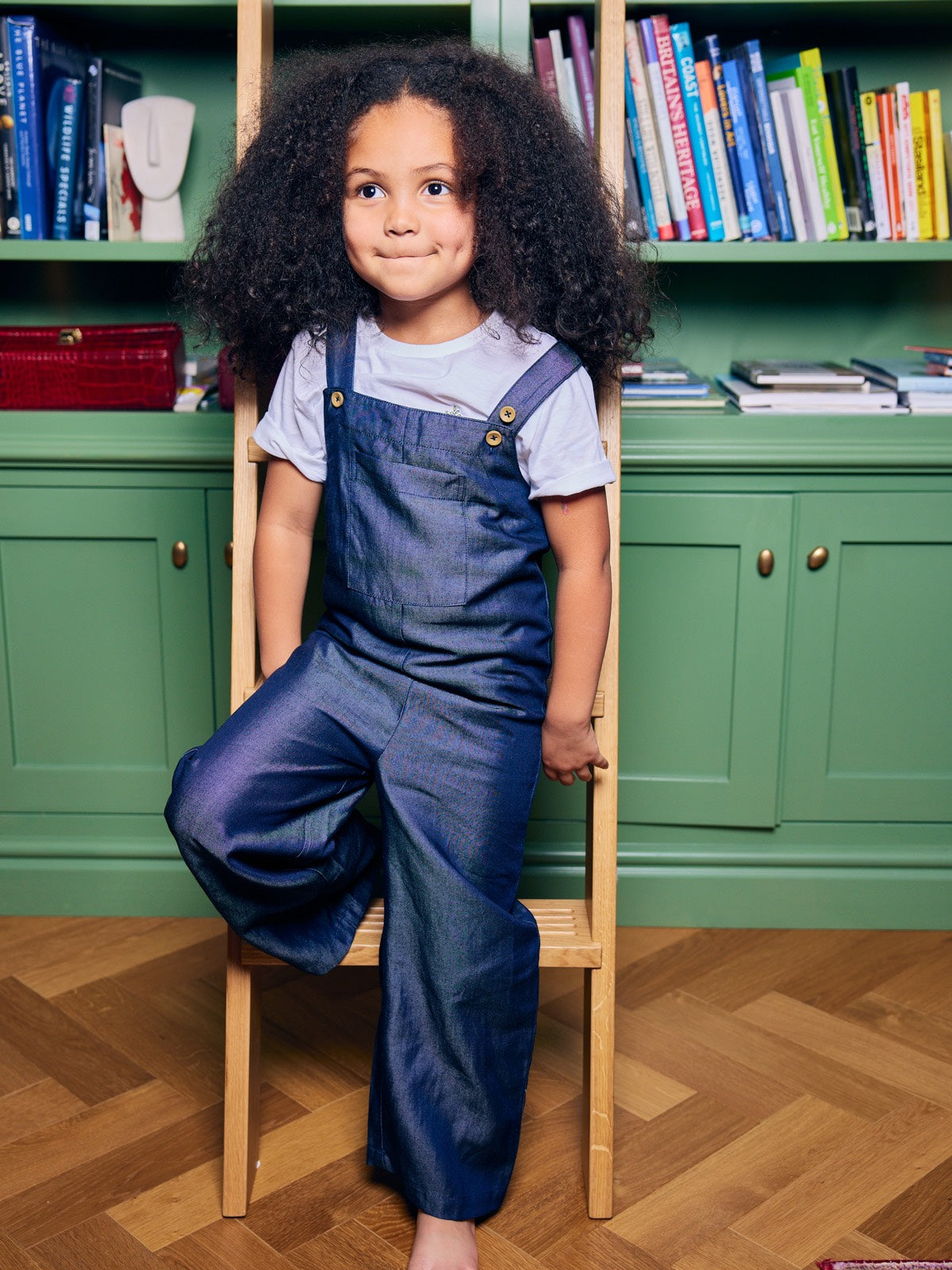 A child wearing the Sofia sustainable kids dungarees, pictured leaning against a ladder in front of a bookshelf.
