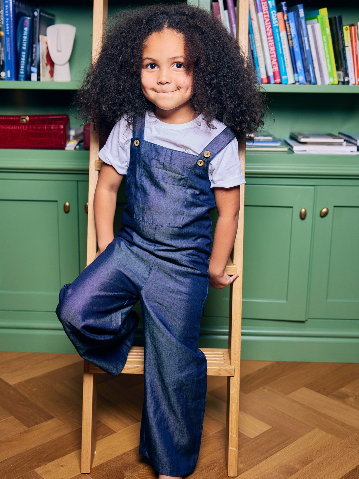 A child wearing the Sofia sustainable kids dungarees, pictured leaning against a ladder in front of a bookshelf and smiling at the camera. 