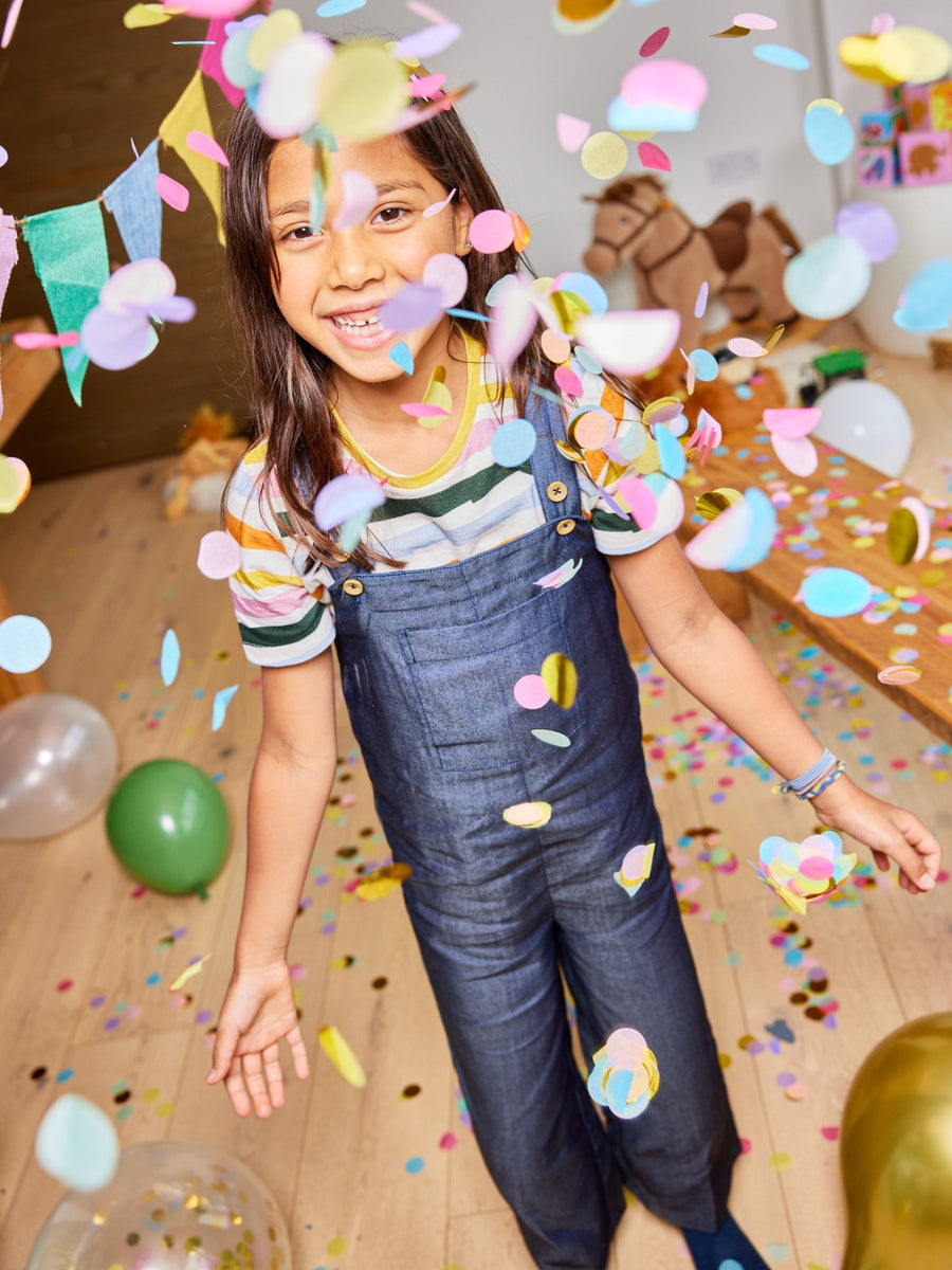 A child wearing the Sofia sustainable kids dungarees, pictured with falling confetti and balloons and smiling at the camera.