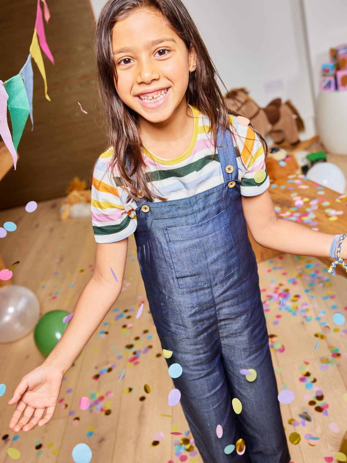 A child wearing the Sofia sustainable kids dungarees, pictured with falling confetti and balloons and smiling at the camera.