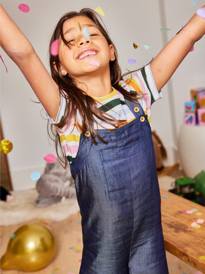 A child wearing the Sofia sustainable kids dungarees, pictured with falling confetti and balloons and smiling at the camera.