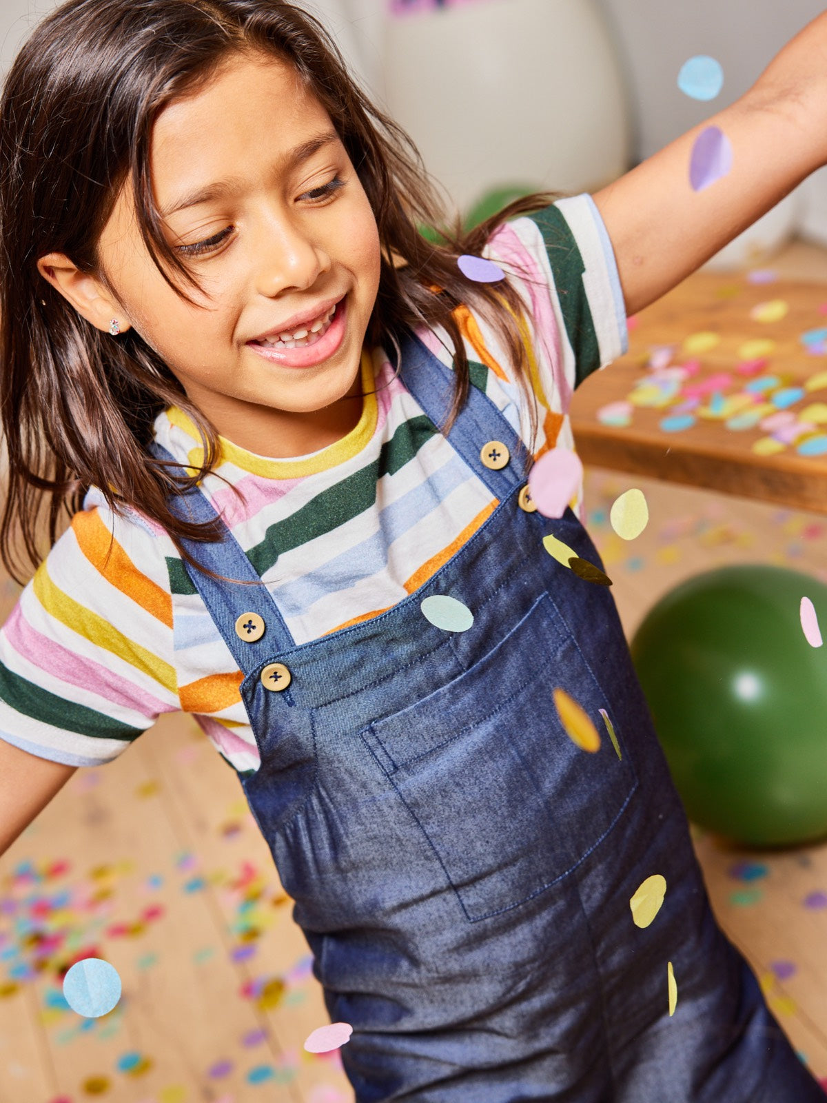 A child wearing the Sofia eco-friendly kids dungarees, pictured with falling confetti and balloons.