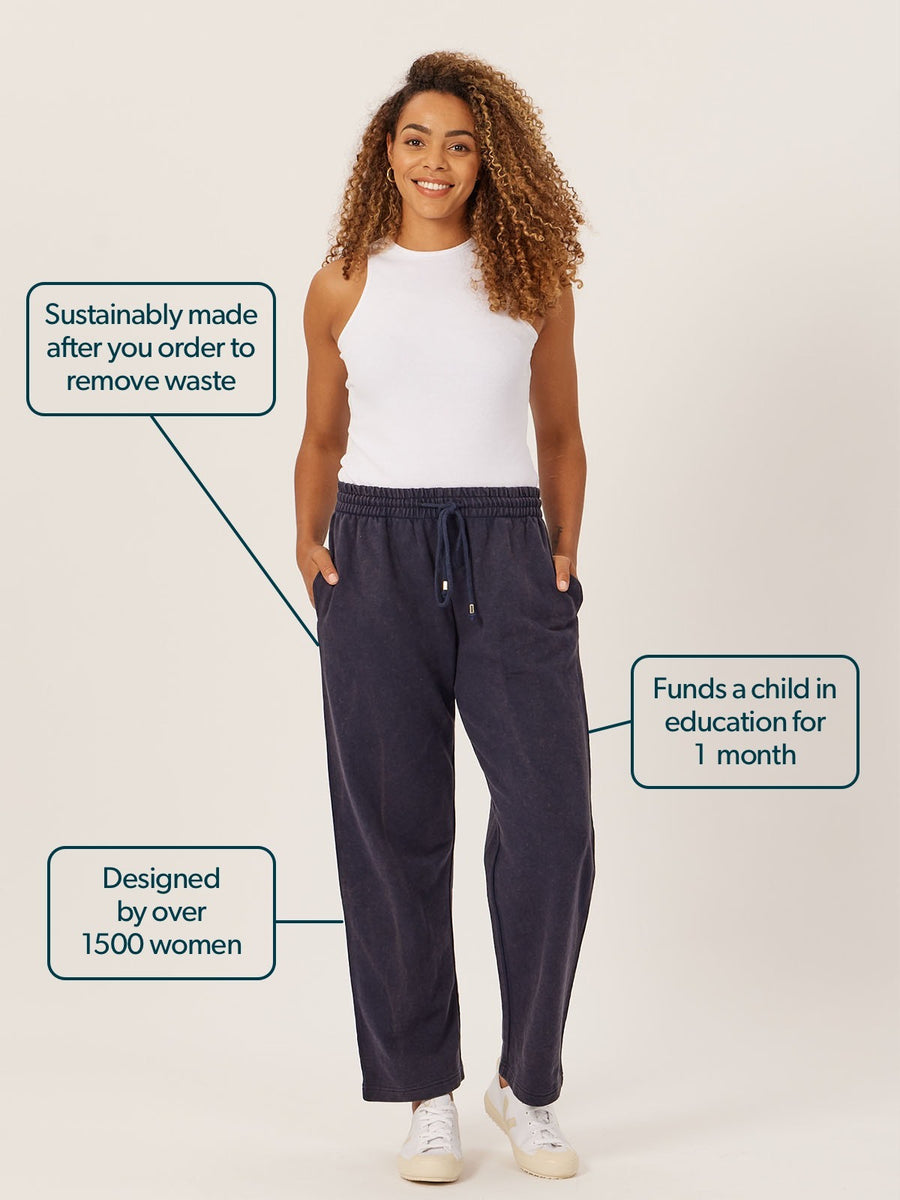 Blue stonewash sustainable joggers from This is Unfolded, worn with a white vest top. Three speech bubbles with text come off the image, saying: 1. Sustainably made after you order to remove waste. 2. Funds a child in education for 1 month. 3. Designed by over 1500 women.