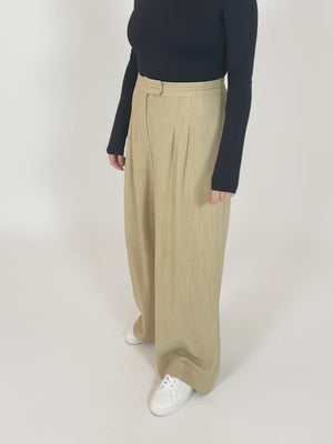 A video of a model wearing the Lana trousers in sand and a long sleeve black top. The model is turning around to display the trousers, and  is placing their hands in their pockets. 